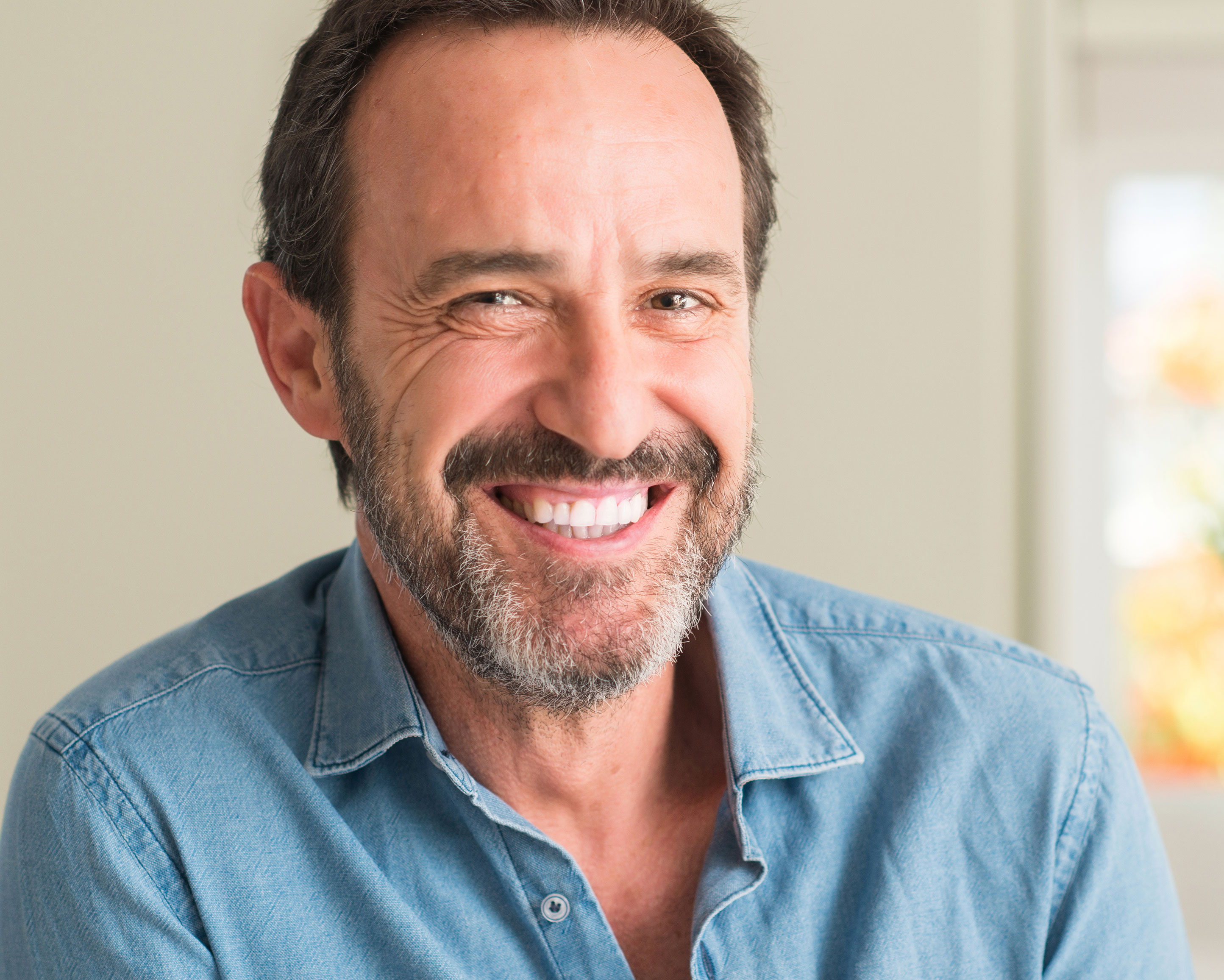 The image displays a man with a beard smiling at the camera, wearing a denim shirt and standing against a backdrop that appears to be an interior wall.