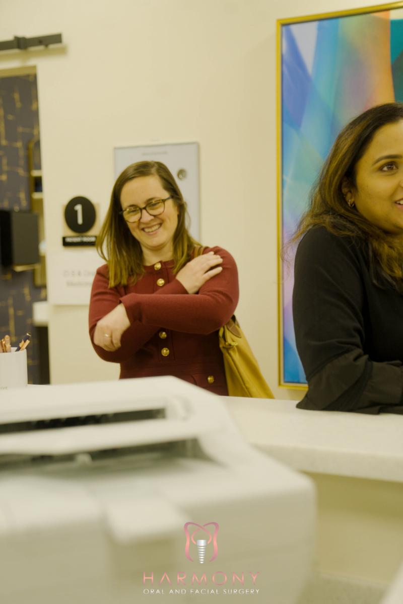 A photograph of two individuals inside an office setting one woman with her arms crossed smiles at the camera while another person, partially visible, stands behind a counter.