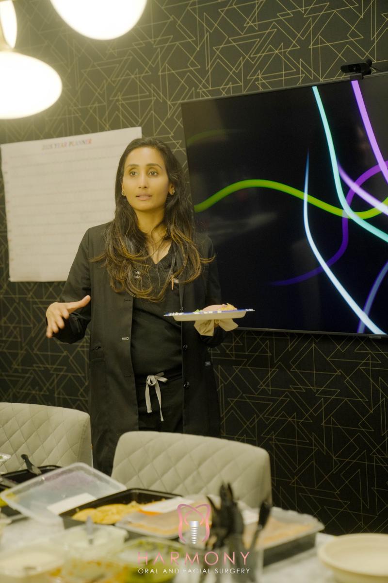 Woman standing at podium with presentation screen behind her, gesturing towards camera.
