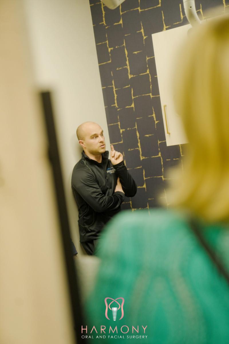 The image shows a man standing in front of a mirror, with another person partially visible on the left side of the frame. They are indoors, and the setting appears to be a room with a patterned wall.