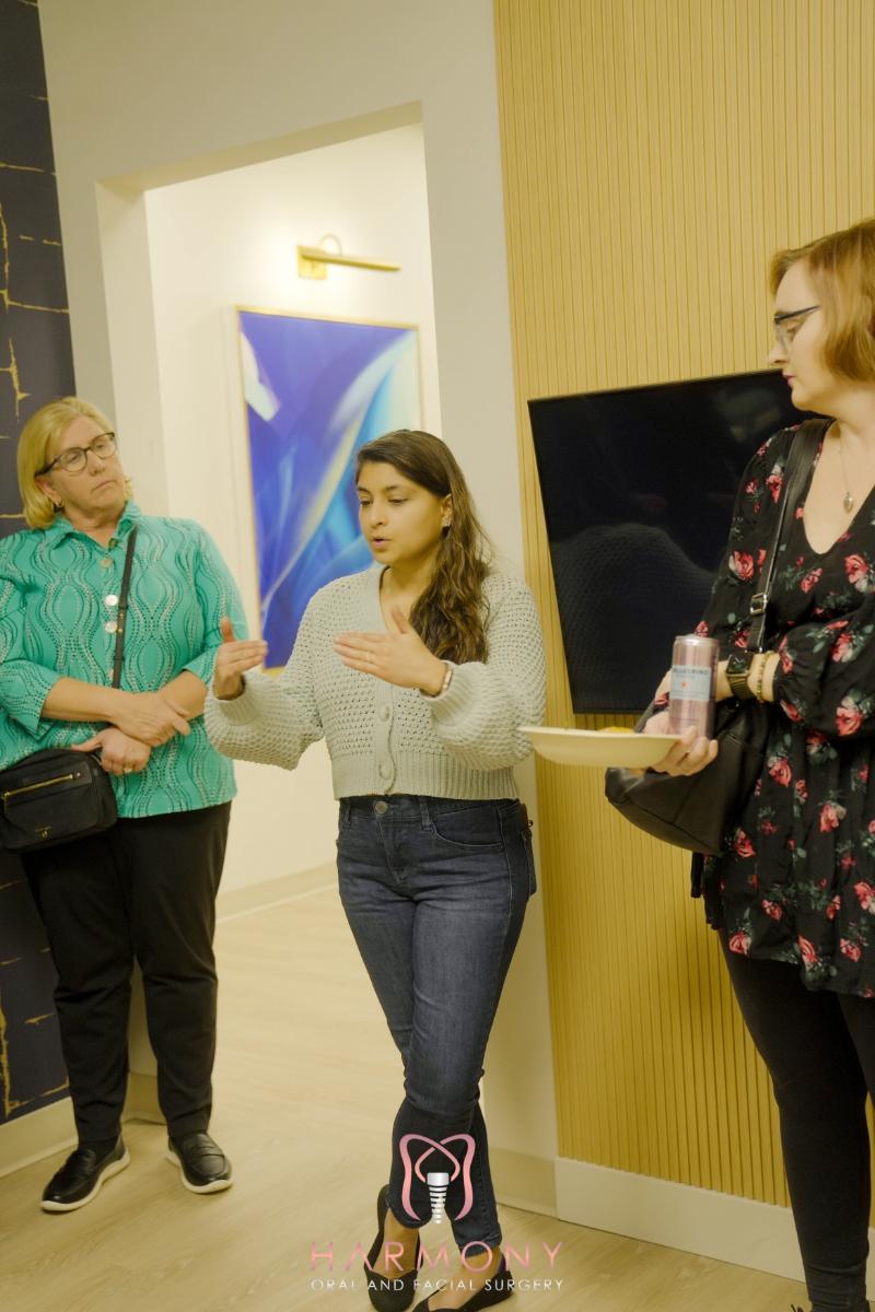 In the image, there are three individuals standing in an indoor setting with a modern decor. A woman in the center is gesturing with her hands while speaking, and two other people are observing her. The room has contemporary furnishings, including a wall-mounted light fixture and a sleek, dark-colored floor lamp.