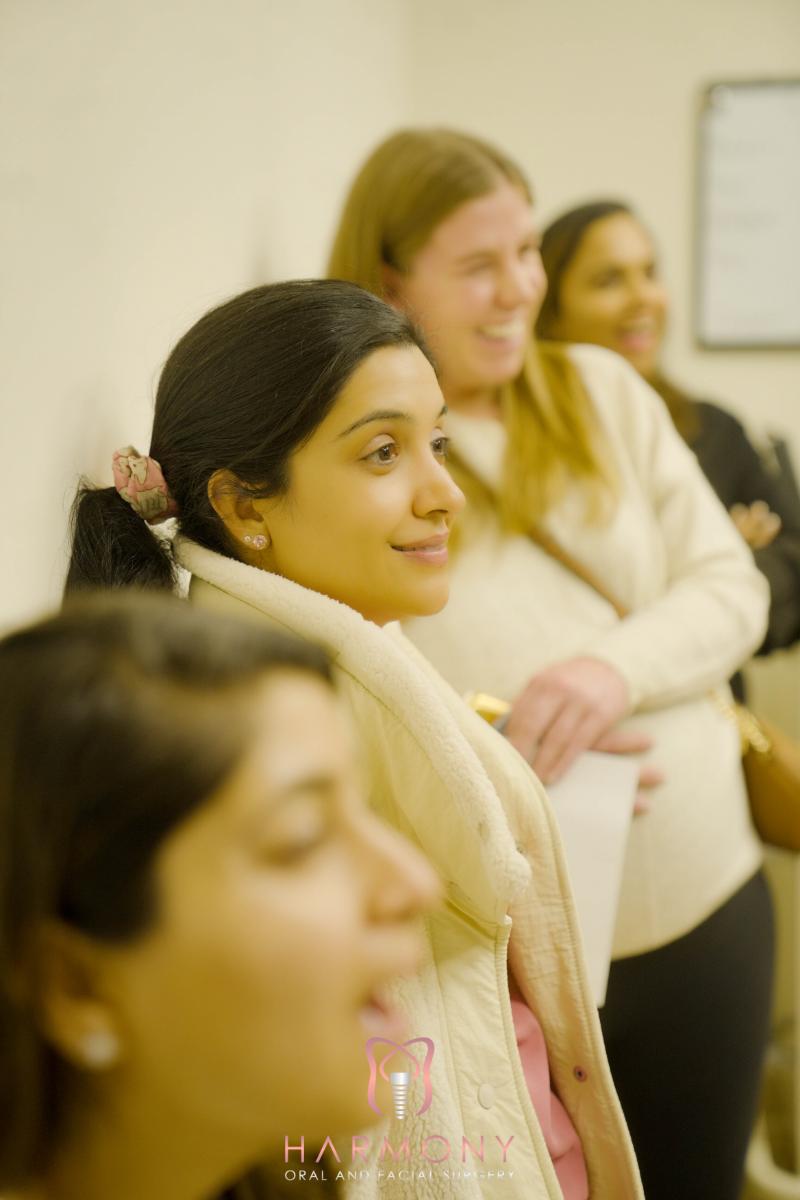 The image shows a person seated with others, smiling at the camera, wearing a scarf and a jacket, in an indoor setting that appears to be a meeting or event.
