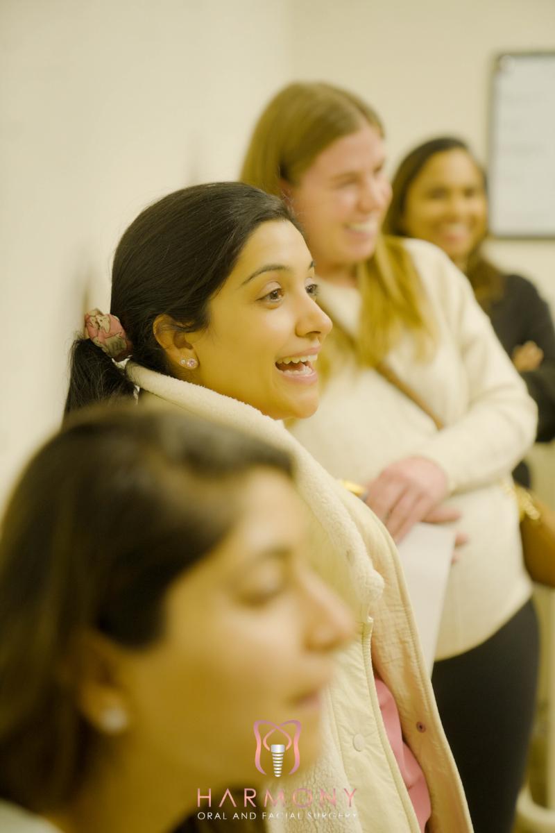 The image shows a group of four people standing together, with two women smiling at the camera and two others looking away, in an indoor setting that appears to be a classroom or lecture hall.