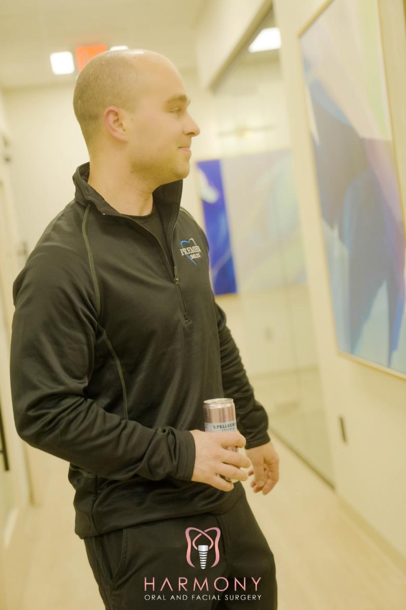 The image shows a man standing in an indoor setting with a beverage in his hand, wearing athletic attire and a lanyard, looking towards the camera.