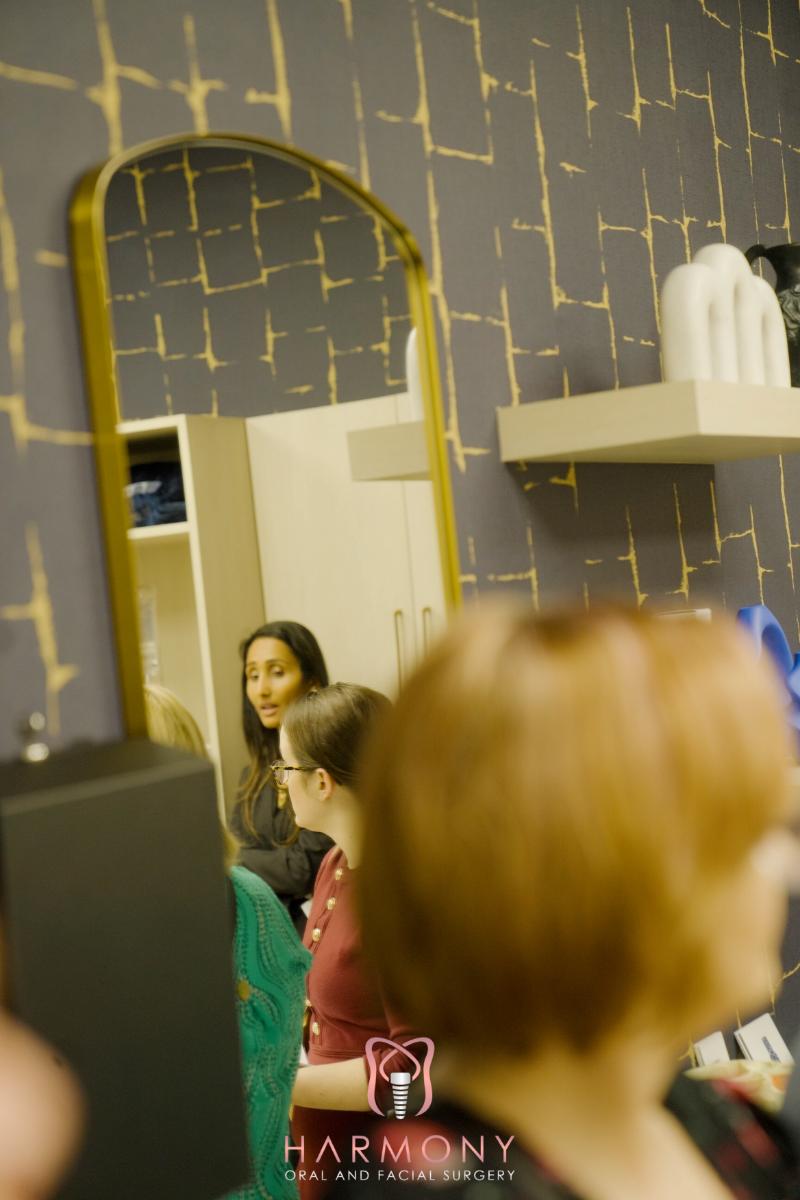 The image shows an interior space with individuals present, featuring a large mirror reflecting part of the room, a wall with a geometric pattern, and various items such as bottles and a handbag.