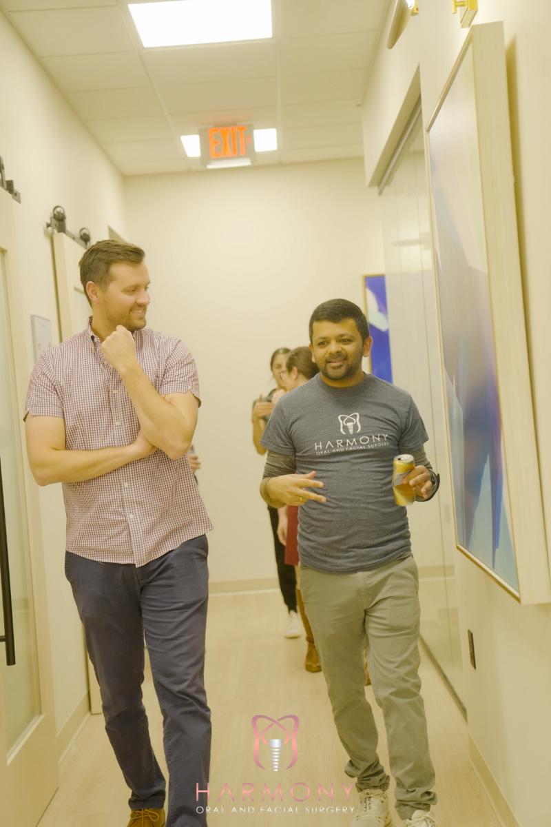 Two men walking down a hallway, one holding a drink, both smiling and engaged in conversation.