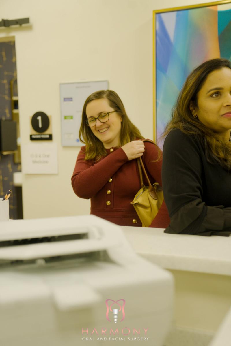 A woman standing behind a counter with a laptop, smiling at the camera, wearing glasses and a red top.