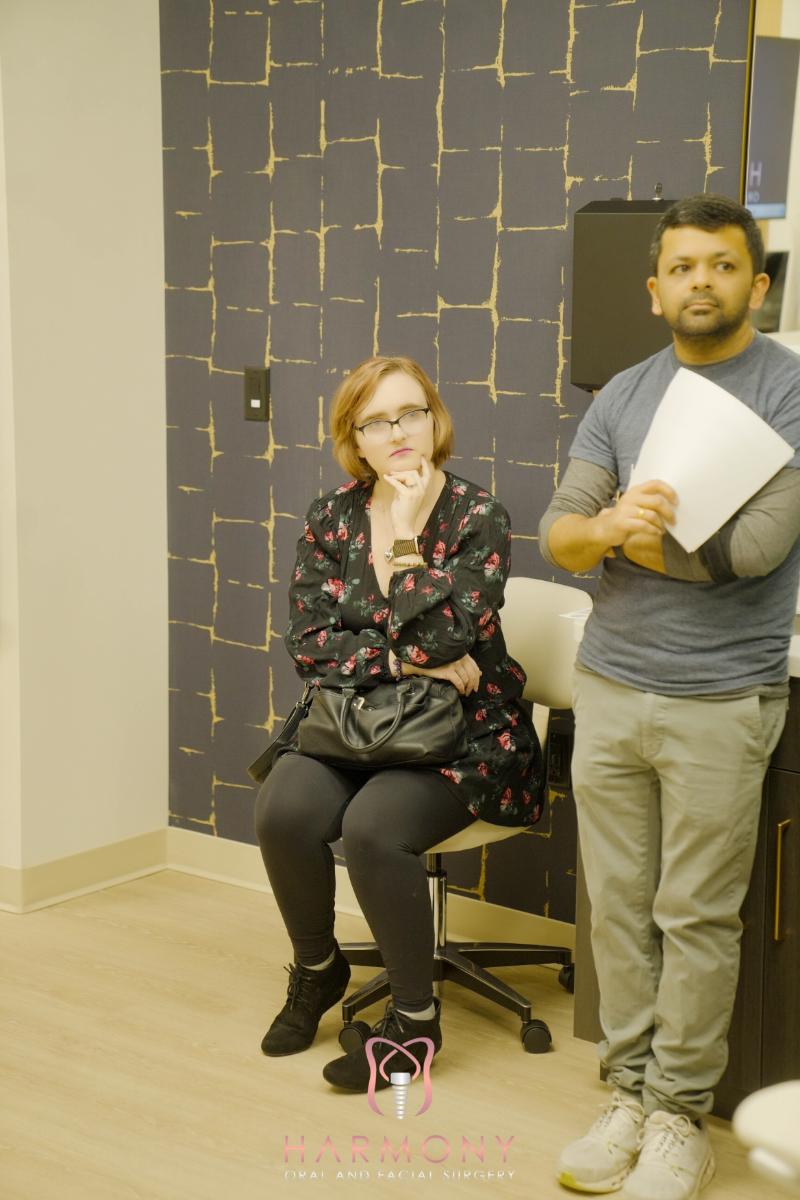 The image shows a man standing next to a woman seated on a chair, both appear to be engaged in a conversation. They are indoors, possibly in an office setting, with a decorative wall behind them.