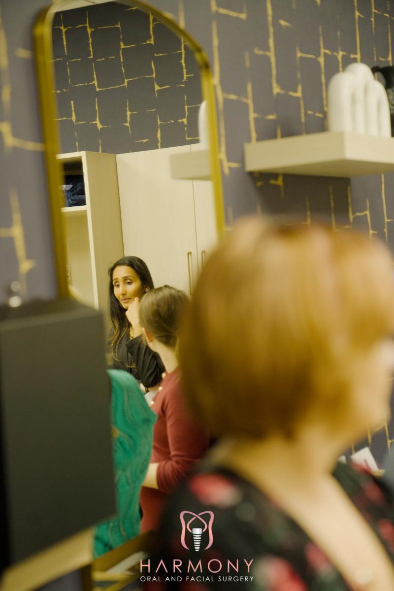 The image shows a woman sitting at a salon station with her back to the camera, facing a mirror where another person's reflection can be seen.
