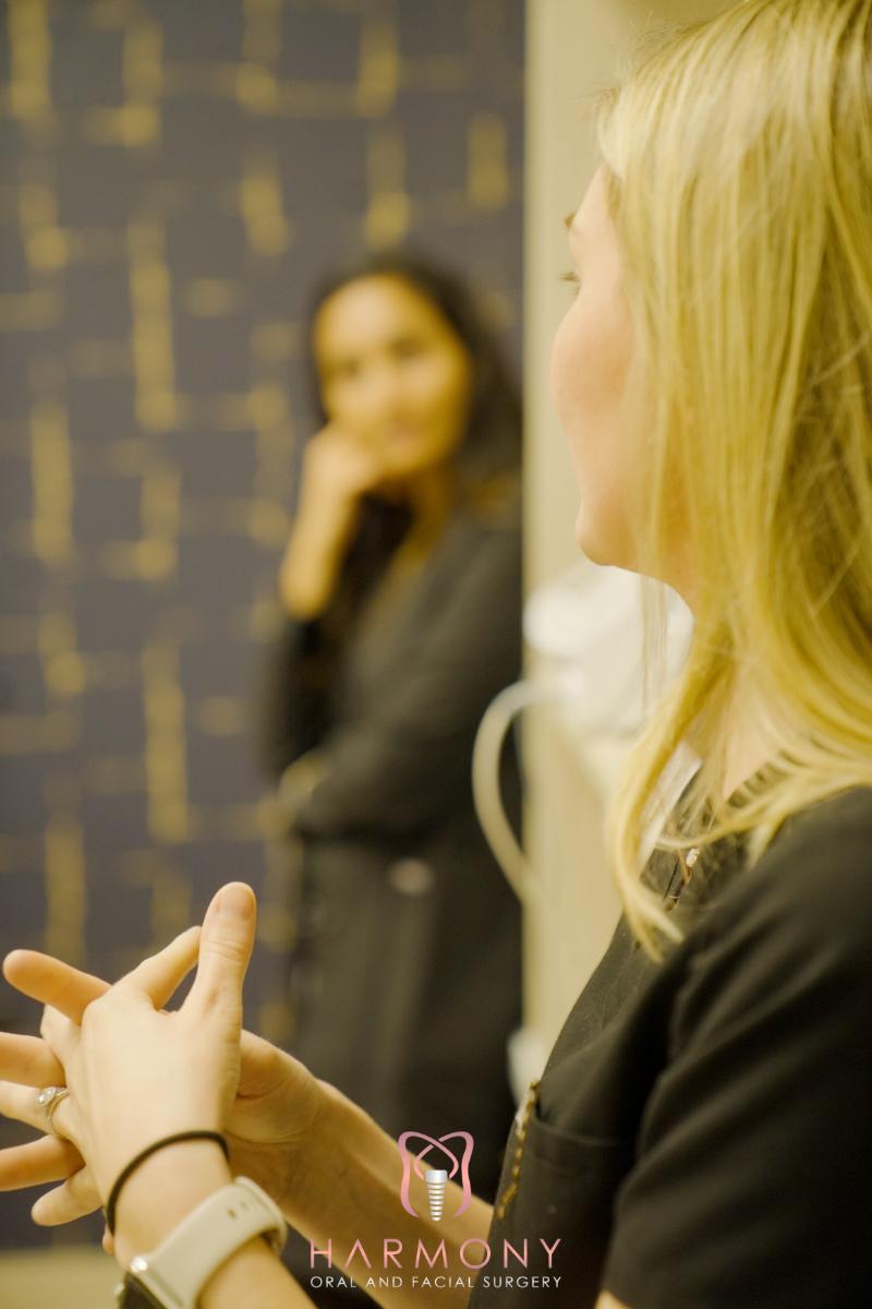 The image features a woman standing at a podium with her hands clasped together, appearing to be speaking or presenting, alongside another woman who is attentively observing her.