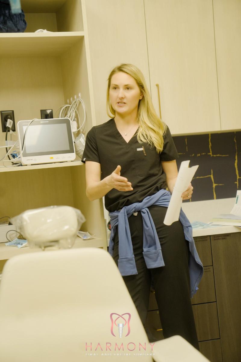 A woman in a blue shirt stands in front of a dental chair, gesturing with her right hand while addressing an audience.