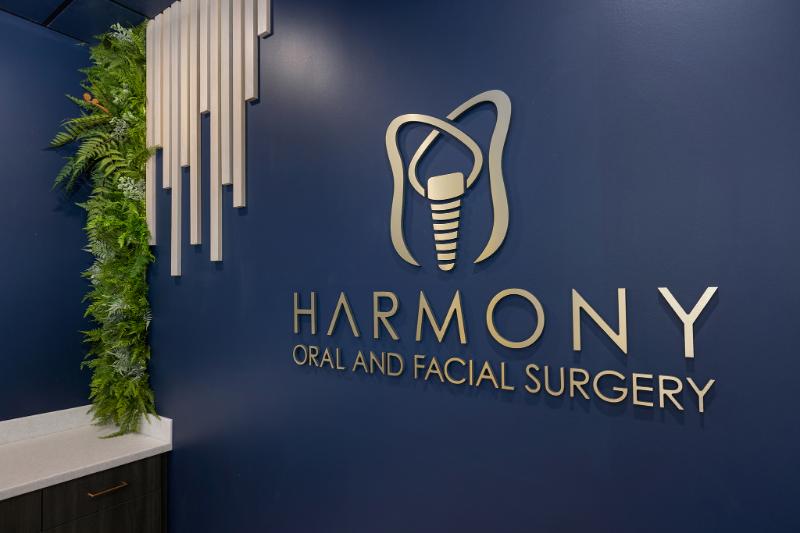 The image shows an interior space with a sign that reads Harmony Oral Surgery featuring a logo that includes a stylized letter E inside a circle, set against a blue background with a minimalist design and modern typography.