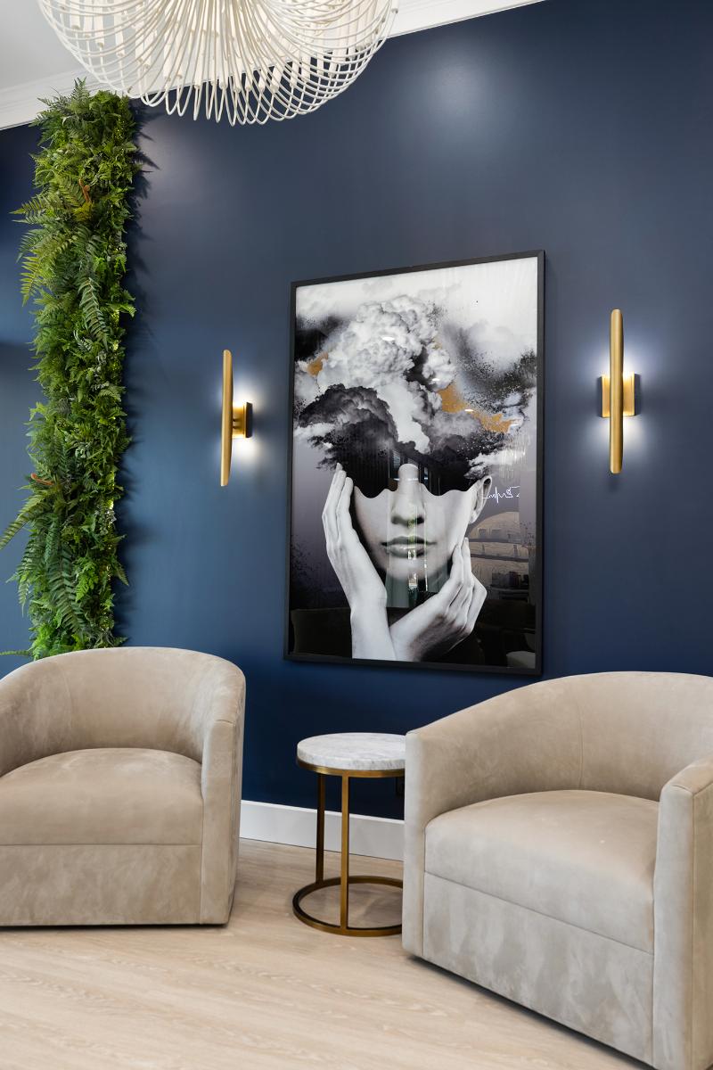 A modern living room with blue walls, a large framed picture on the wall, two armchairs, a coffee table, and a chandelier hanging from the ceiling.