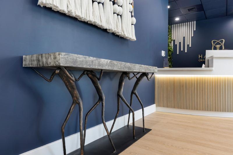 The image shows a room with a blue wall, a long wooden table with three legs at the center, a white countertop, a plant on the floor, and a modern interior design style.