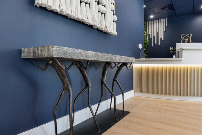 The image shows a modern interior space with a dark blue wall, a light-colored wooden floor, a minimalist console table with two vertical elements resembling legs, and a long, rectangular countertop with a metal frame. There are three abstract sculptures of varying heights on the countertop, and a large abstract piece hanging above it. The space is well-lit and has a contemporary feel.