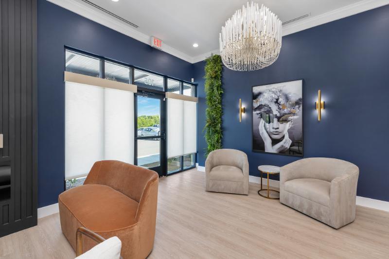 The image shows a modern interior space with blue walls, a chandelier hanging from the ceiling, a large window allowing natural light into the room, a comfortable seating area with a couch and chairs, and a large painting on the wall.