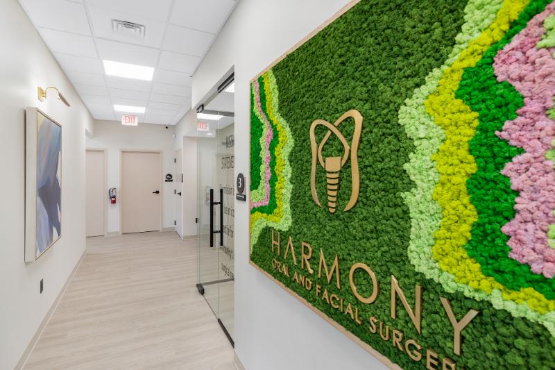 A photograph of a modern interior space featuring a large, colorful wall mural with a floral pattern and text, including Harmony and Surgery, along with a logo resembling a stylized letter M within a shield-like emblem, set against a white background.