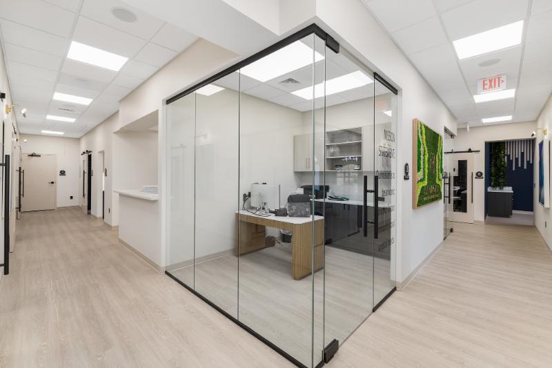 The image shows an interior space that appears to be a dental office with a modern design, featuring a large glass wall separating the waiting area from the treatment rooms, and the room is well-lit with natural light coming through the windows.