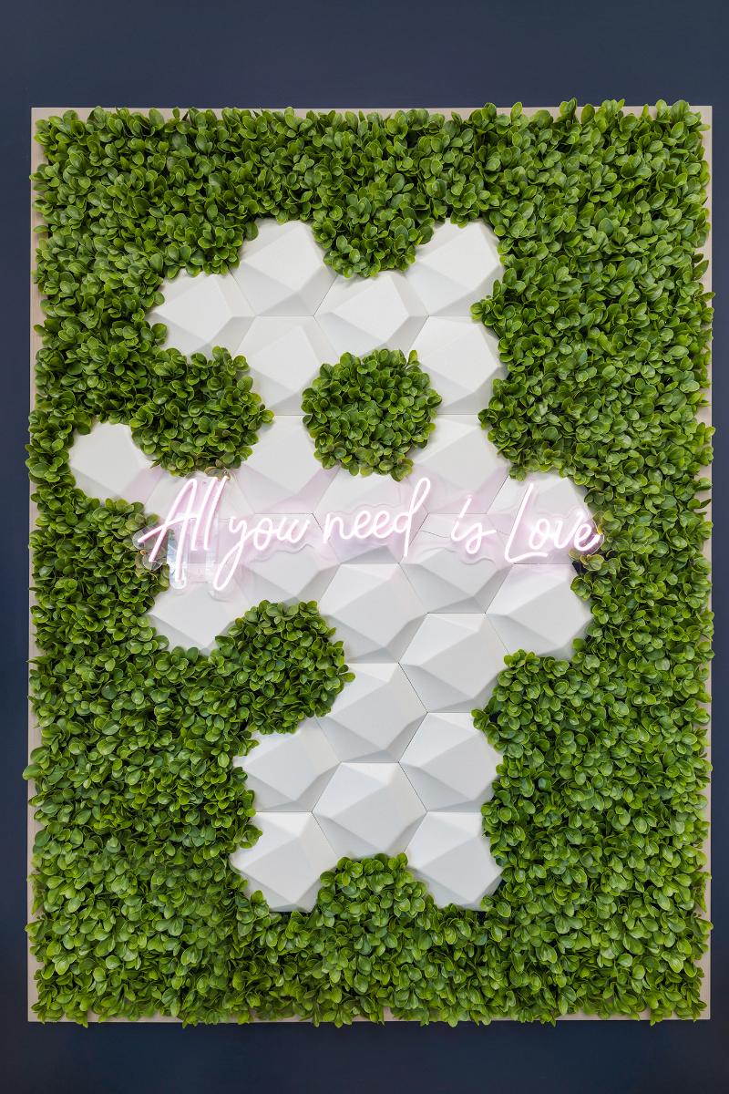 The image shows a three-dimensional wall decoration with a green plant base, featuring a large white sculpture resembling a person with their arms outstretched and a pink text overlay that says 'ALL YOU NEED IS LOVE.