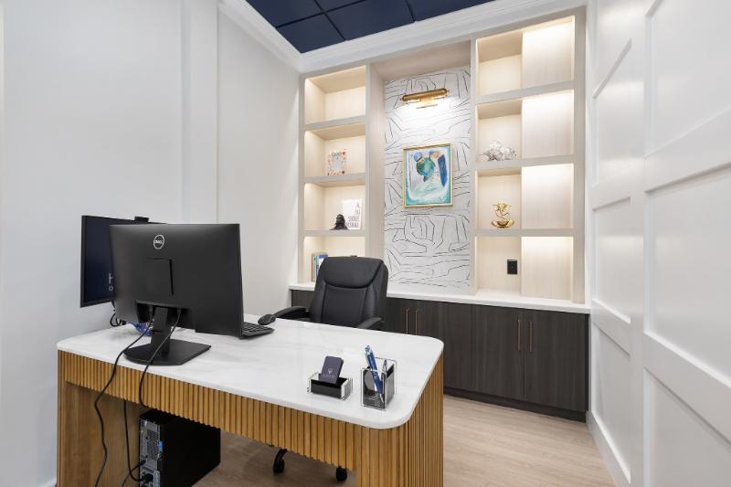 The image shows an interior view of a modern office space with a desk, chair, shelves, and a television screen.