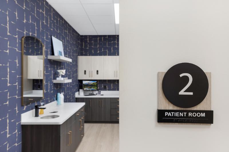 The image shows a modern interior space with a sign indicating 'Patient Room 2' on a wall, featuring a number 2 and a patient room symbol, within an environment that includes a desk, cabinets, and various medical-related items.