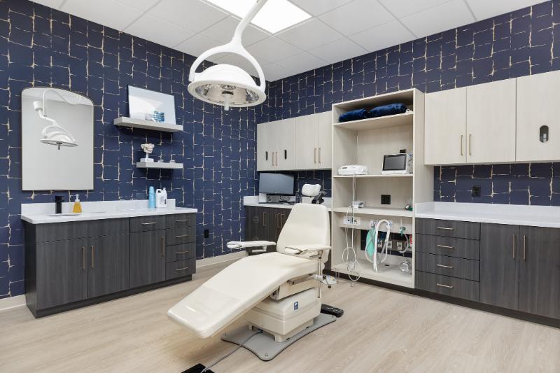 The image shows an interior view of a dental office with modern furnishings, including a reception area, dental chair, examination room, and equipment.