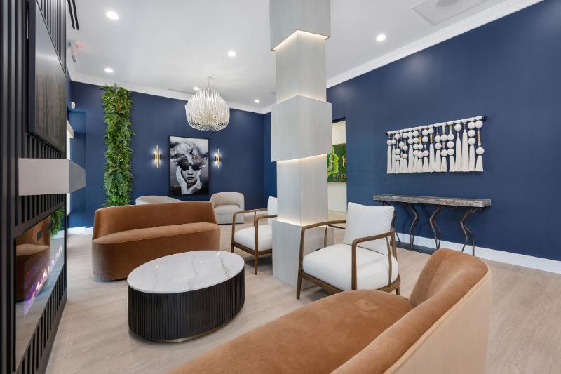 This is a photograph of an interior space with modern furniture and design elements, featuring a living room area with seating options, a dining table, and a wall-mounted painting, and a separate sitting area with a couch and additional seating. The color palette includes shades of blue, brown, and white, and the room has a contemporary feel.