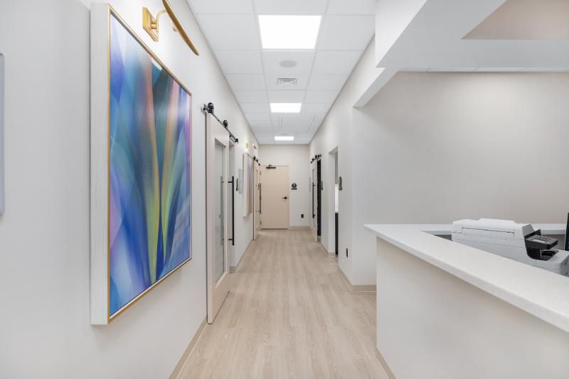 The image shows an interior view of a modern medical office with white walls, a clean and well-lit environment, and a hallway leading to various rooms including a waiting area and examination rooms.