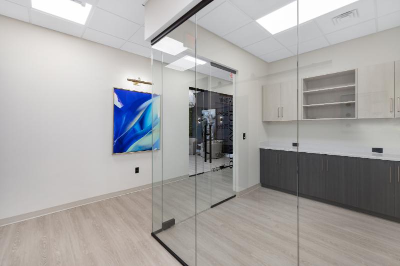 The image shows a modern interior space featuring a glass-walled room with a sleek design, including a kitchen area with cabinets and countertops, a wall-mounted TV, and a window with a view of a blue sky.
