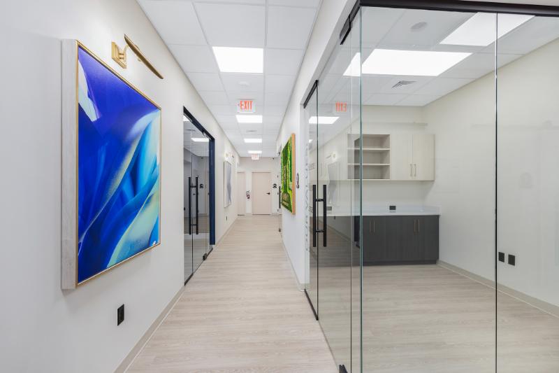 The image depicts a modern office interior with a long hallway leading to a reception area featuring a large glass partition, a white ceiling, and framed artwork on the walls.