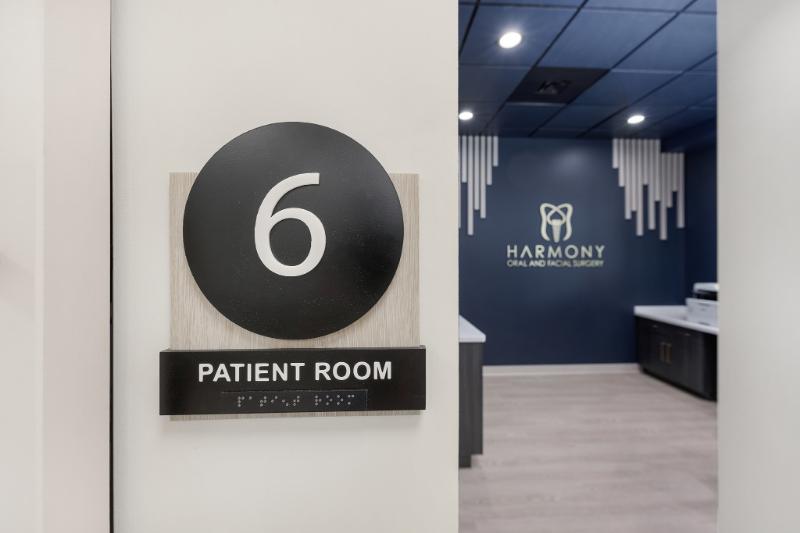 The image shows an interior space with a sign indicating room number 6, which suggests it is part of a larger facility such as a hotel or hospitality establishment. The sign has a modern design with a white background and black text, featuring a logo that appears to be a stylized letter 'H'. There's a sign on the wall behind the door that reads 'Patient Room' in capital letters, indicating that this is a medical facility. The room itself is not visible in full but seems to have a clean and professional appearance with blue walls and white trim.