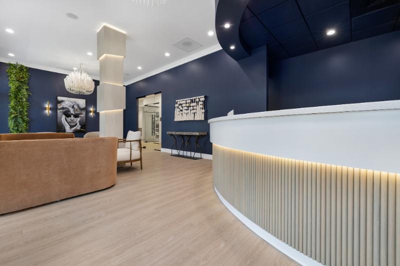 The image shows an interior space with a modern design, featuring a reception area with a curved counter, seating, and decorative elements such as plants and artwork on the walls.