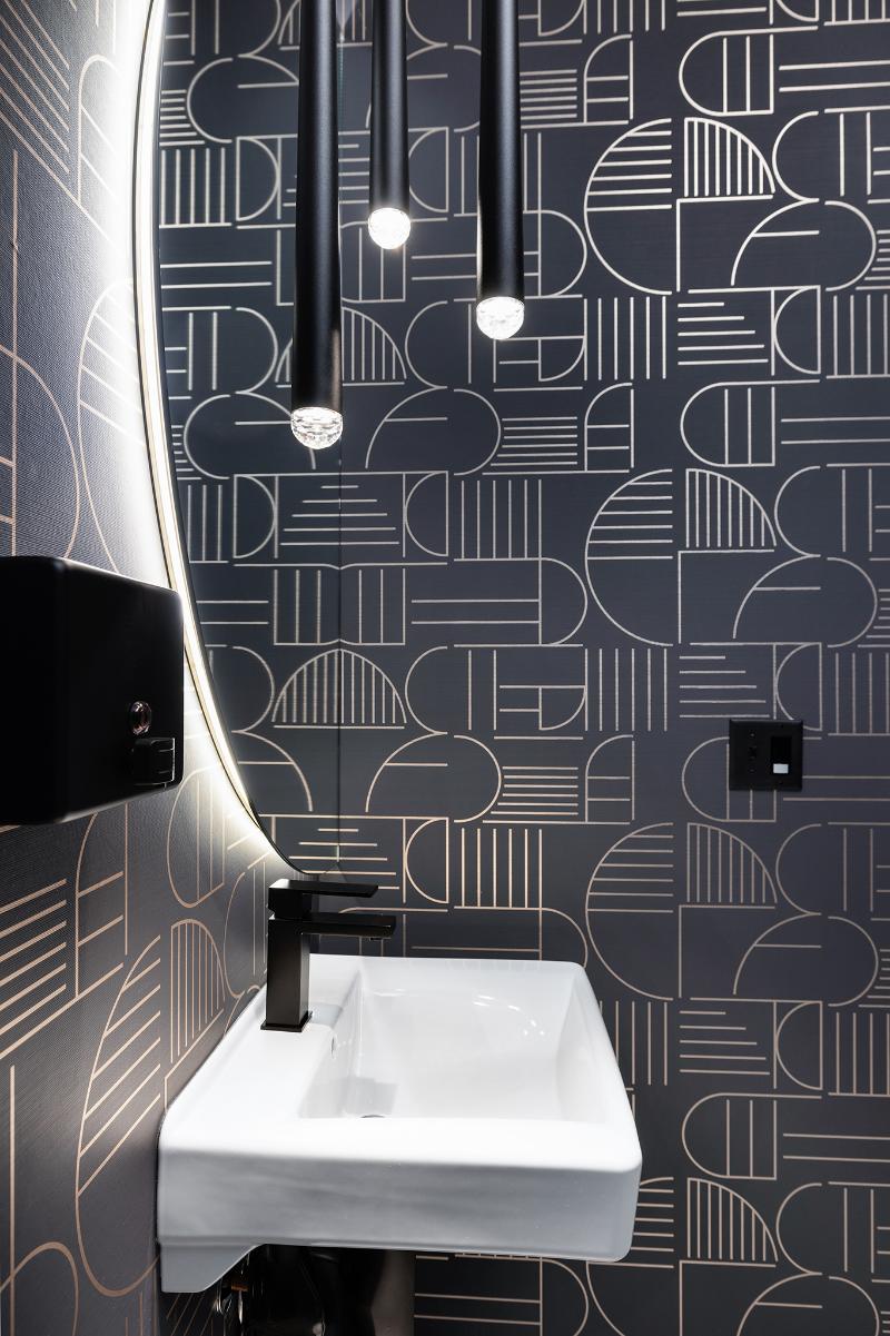 A modern bathroom with a black and white geometric wallpaper design, featuring a sink with gold accents and a circular mirror above it.