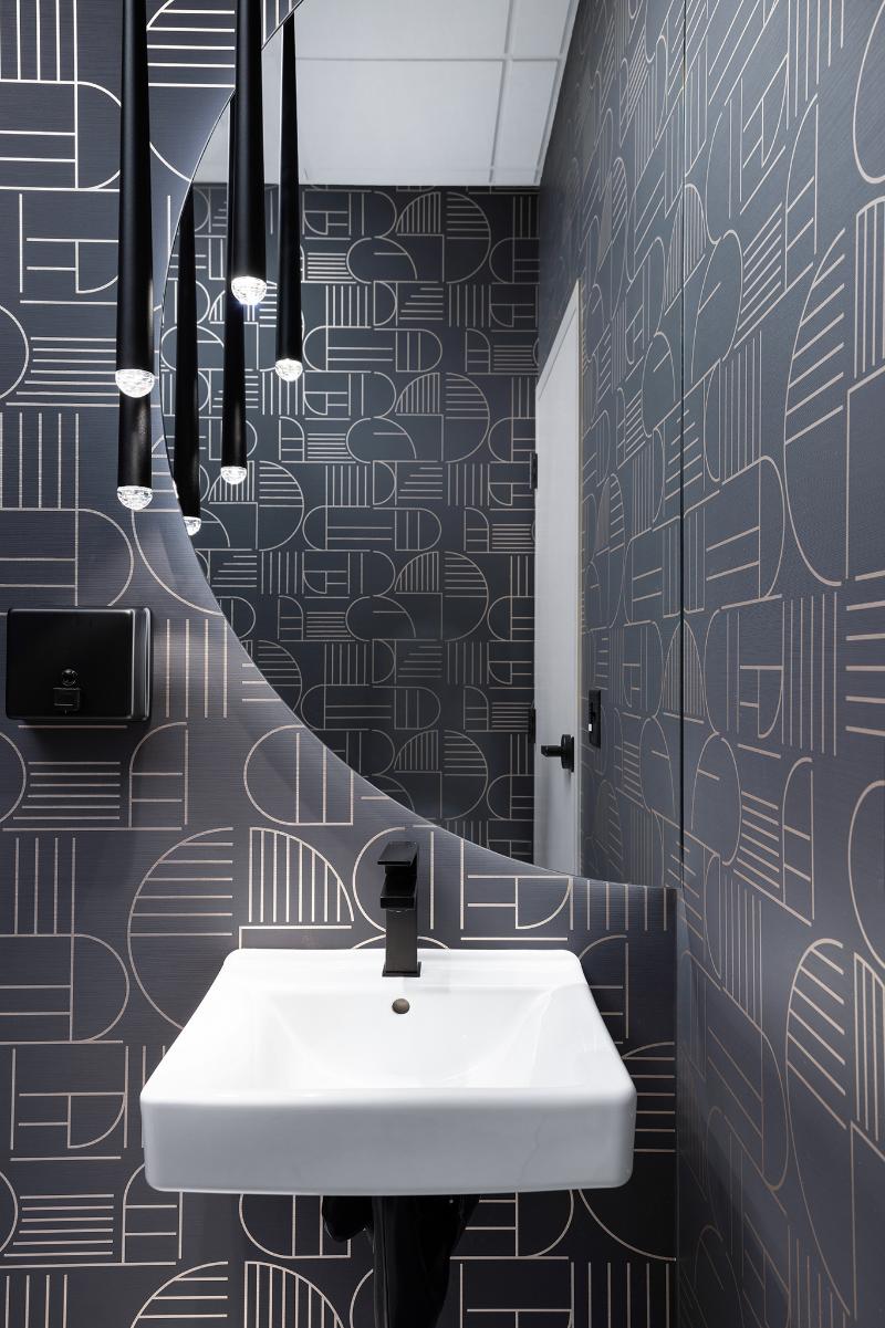The image shows a modern bathroom with a dark color scheme featuring a black and white geometric wallpaper, a sink with a mirror above it, and a patterned backsplash.