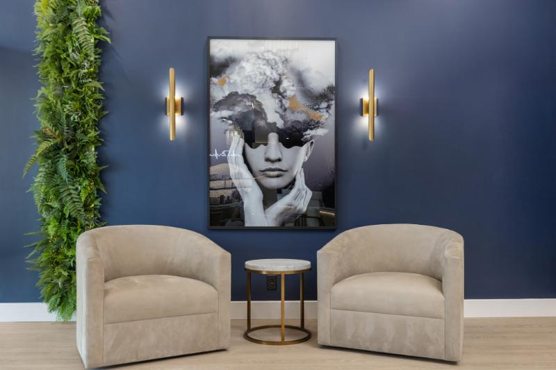 A modern living room setting with a blue wall, framed artwork featuring a person's face, a large potted plant, and a seating area with two chairs and a sofa.