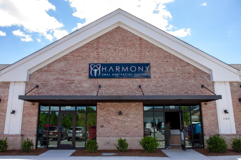 The image shows a building with a sign that reads 'Harmony Dental' outside, indicating it's a dental clinic, with a clear sky and trees visible in the background.