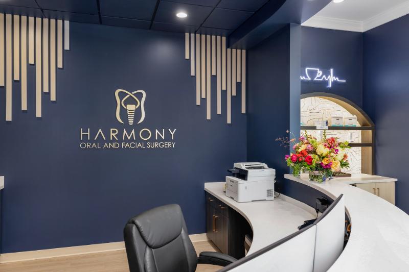 The image shows an interior space with a wall-mounted sign that reads 'Harmony Dental and Facial Spa,' featuring a logo of a toothbrush and toothpaste tube, along with a stylized dental implant graphic, set against a blue background with decorative wooden elements.